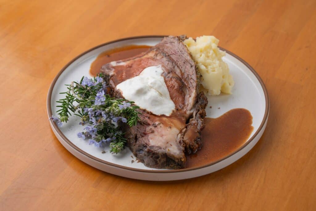 Prime rib served with gravy, mashed potatoes, and herbs on a white plate.